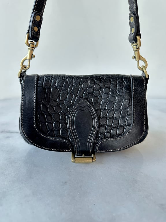Sort skuldertaske i læder med krokodillemønster og messingbeslag, placeret på marmorbord.