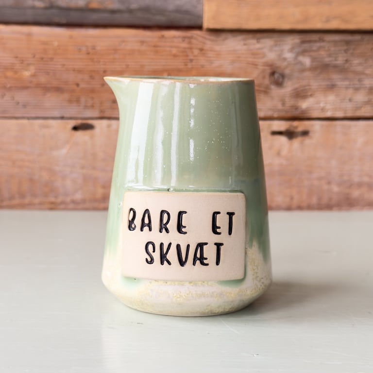 Grøn keramisk mælkekande med teksten "Bare et skvæt" på en beige plade foran, står på et lyst bord med trævæg i baggrunden.