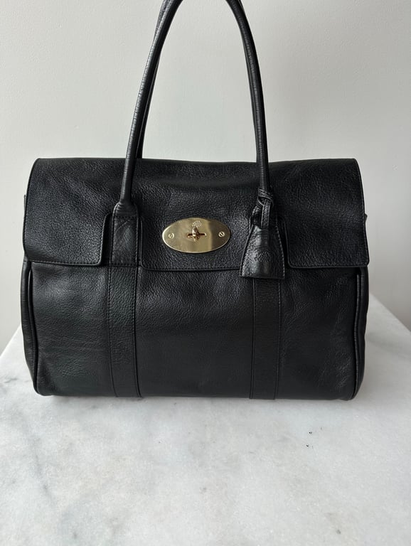 Sort lædertaske med to hanker og guldfarvet lås placeret på en hvid marmorflade foran en lys væg.