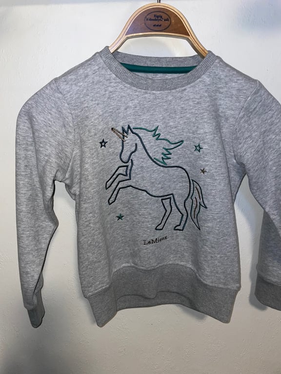 Grå børne-sweatshirt med en enkel tegning af en enhjørning og små stjerner, hængende på en bøjle.