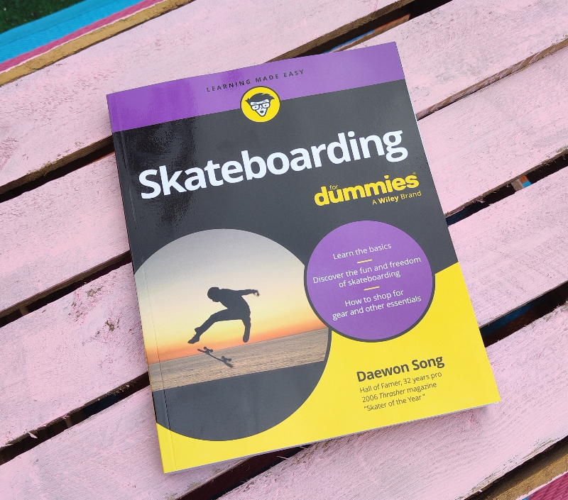 Bog med titlen "Skateboarding for dummies" lagt på en lyserød træbænk, med et billede af en person der laver et skateboard-trick ved solnedgang på forsiden.