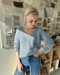 Kvinde iført lyseblå, strikket trøje og blå jeans står foran en væg med forskellige strikkede prøver opsat. (1)