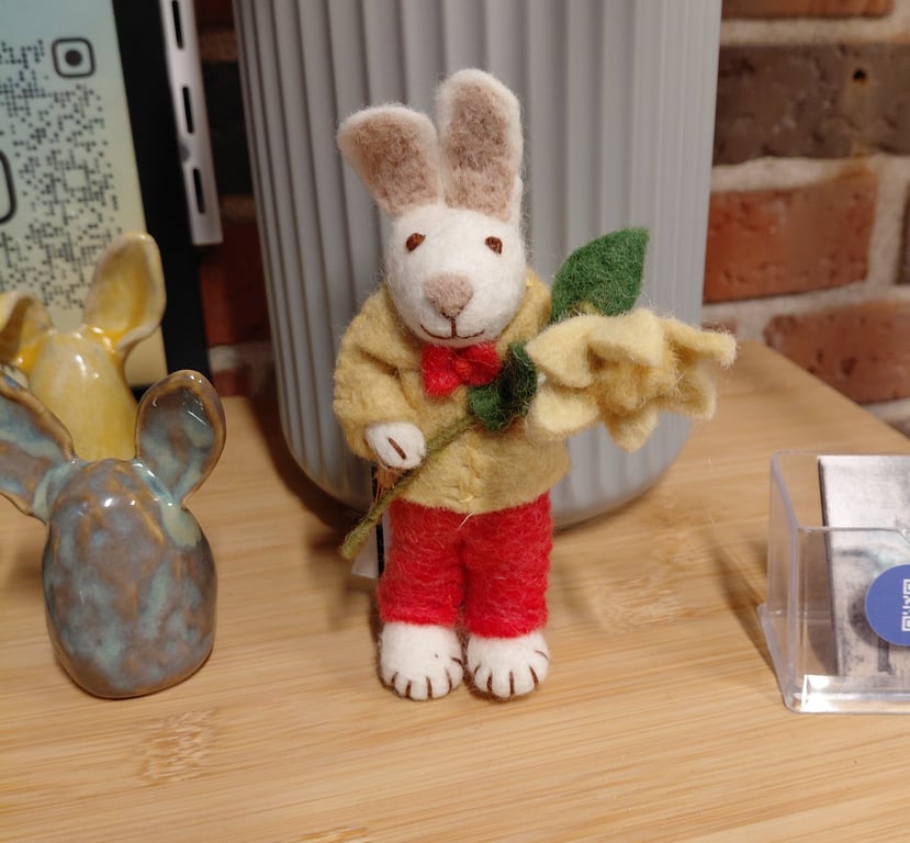 Filtet kaninfigur med gul jakke, røde bukser og rød sløjfe holder en gul blomst på et træbord med keramikkaniner og en grå vase i baggrunden.