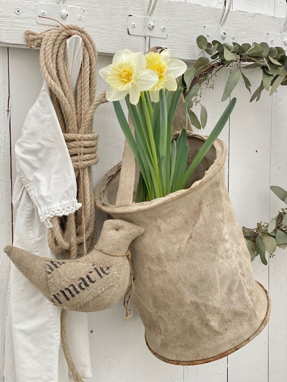 Rustikt ophæng på hvid trævæg med to påskeliljer i et groft lærredsstof, et bundt reb, et hvidt klædestykke og en stoffugl med tekst.