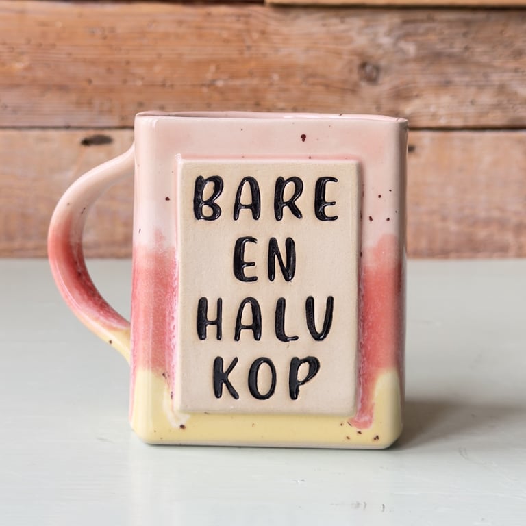 Billede af en firkantet keramikkop i rosa, gul og rød med teksten "BARE EN HALV KOP" skrevet i sort på forsiden.