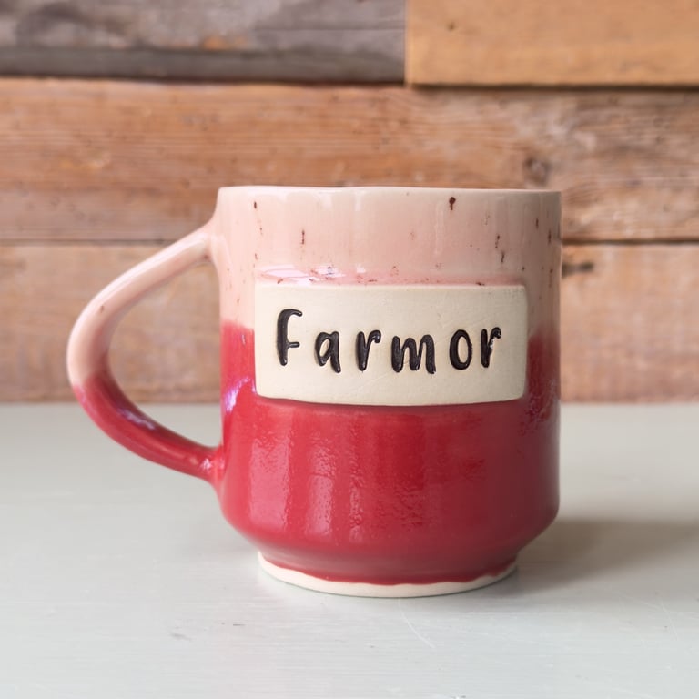Keramikkrus i rød og beige med teksten "Farmor" på forsiden, placeret på et lyst bord med en træbaggrund.