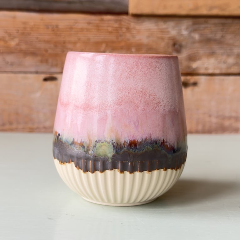 Keramisk vase med flækket beige bund og glaseret overflade i nuancer af pink, sort og grøn, placeret på hvid overflade med træbaggrund.