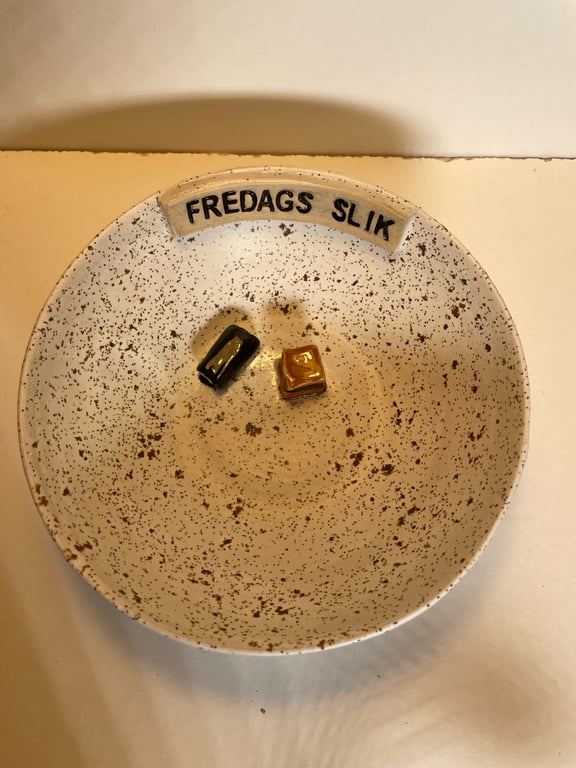 Tallerken med brune prikker og teksten "FREDAGS SLIK" øverst, indeholdende to stykker slik, et sort og et gult.