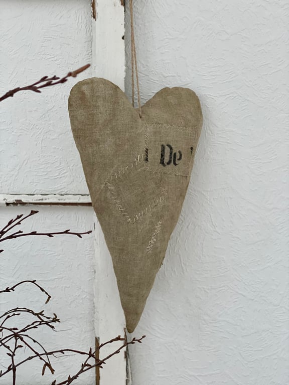 Hængende hjerteformet stofdekoration i beige farve med udsmykkede sting, ophængt på en hvid tekstureret væg.