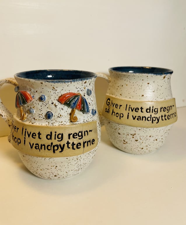 To beige keramikkrus med blå inderside og farverige paraplyer som dekoration. På krusene står der på dansk: "Giver livet dig regn - så hop i vandpytterne."