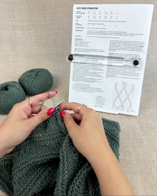 En person strikker med grønt garn foran en opskrift til en vest, der er sat op på en holder. Der er to nøgler af samme grønne garn ved siden af. Personen har røde negle og bærer flere sølvringe. (3)