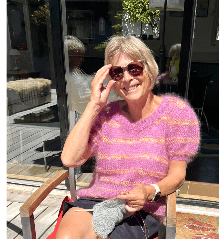 Kvinde iført lilla striktrøje og solbriller, siddende ude på terrasse og strikker.