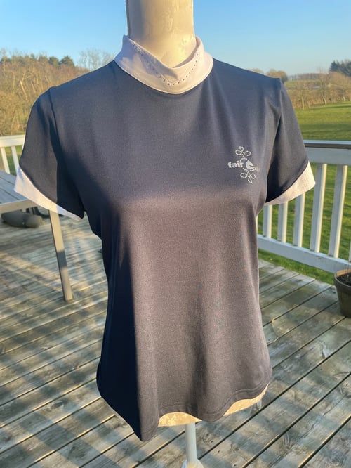 En mørkeblå træningstrøje med korte ærmer og hvide kanter på ærmerne samt en hvid højhalset t-shirt under, vist på en mannequin på en træterrasse med baggrund af græs og træer. (1)