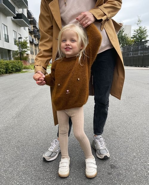 Lille pige iført brun strikbluse med dyremotiv og lyse leggings, der holder hænder med en voksen kvinde iført beige frakke og sorte bukser, ude på en vej i boligområde. (2)