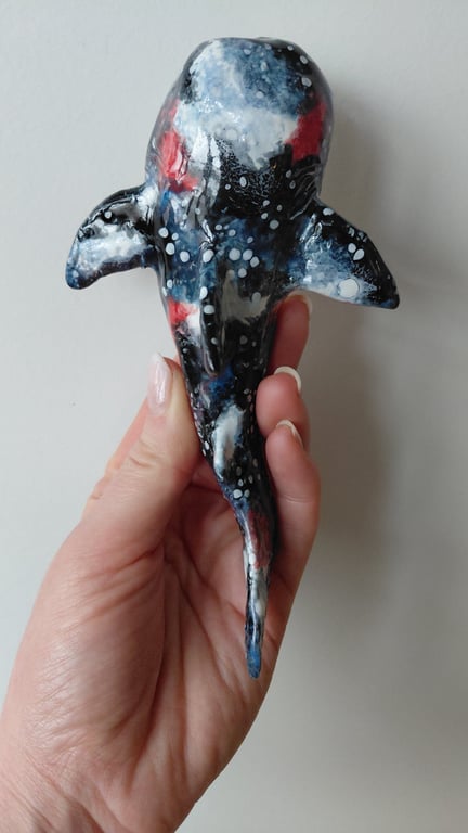 Hånd holder en lille, håndlavet hvalformet figur med en abstrakt, farverig malingsoverflade i sort, hvid, blå og røde nuancer.