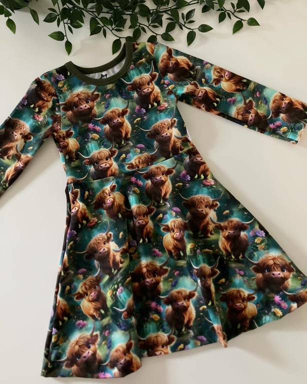 Børnekjole med lange ærmer og mønster af søde højlands-ko-kalve på grøn baggrund, lagt ud på hvid overflade med grønne blade øverst.