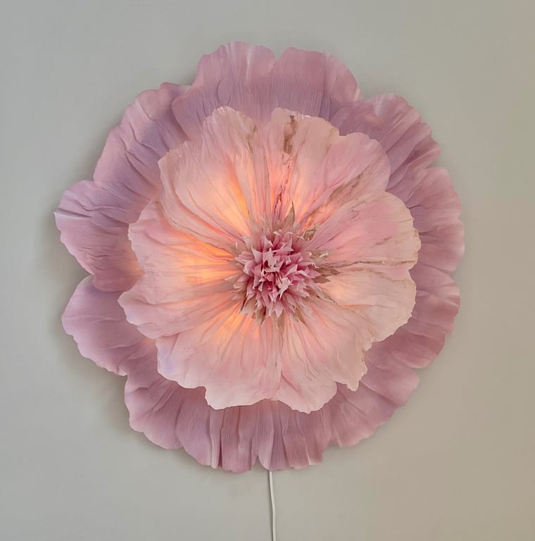 Vægdekoration formet som en stor, lysende blomst i nuancer af lyserød og lilla på en hvid væg.