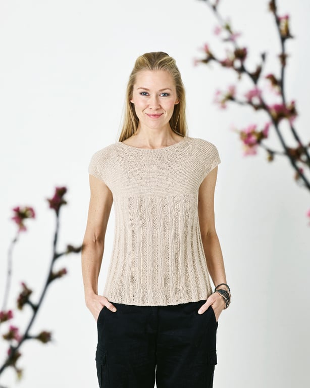 Kvinde iført beige strikket top og sorte bukser står foran en hvid væg med blomstrende grene i baggrunden.