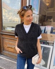 Kvinde iført sort strikket vest over hvid T-shirt, blå jeans og med solbriller på hovedet, står foran en butiksvindue på en byvej. (1)