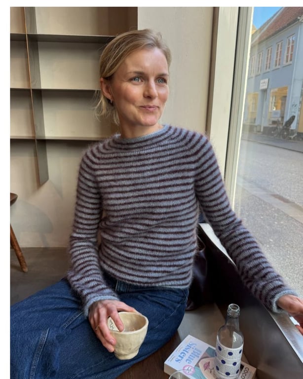 Kvinde iført stribet trøje og jeans, sidder ved vindue med en kop i hånden, indendørs cafémiljø.