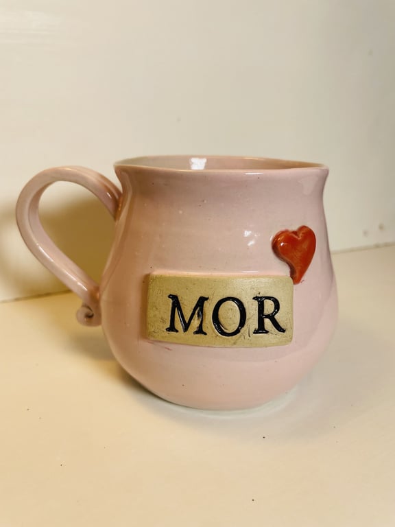 Lyserød keramisk kop med hank, dekoreret med en rektangulær plade med teksten "MOR" og et rødt hjerte ved siden af.