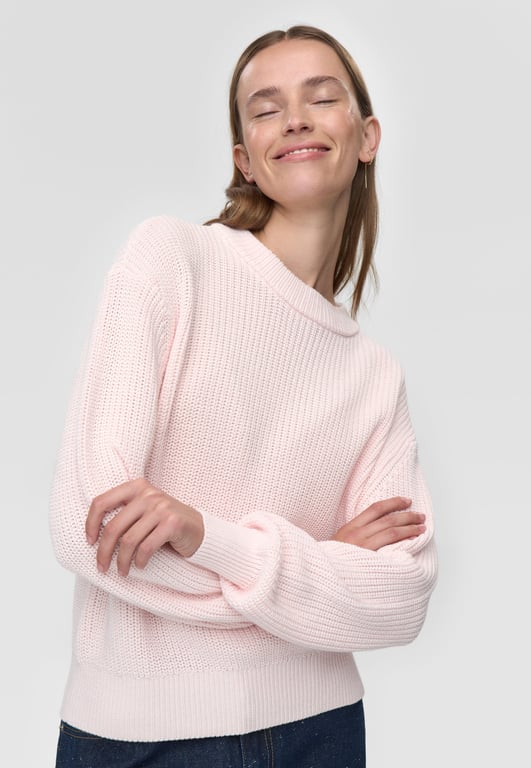 Kvinde iført lys pink, strikket sweater med ærmerne krydset og med lukkede øjne og smilende ansigtsudtryk.