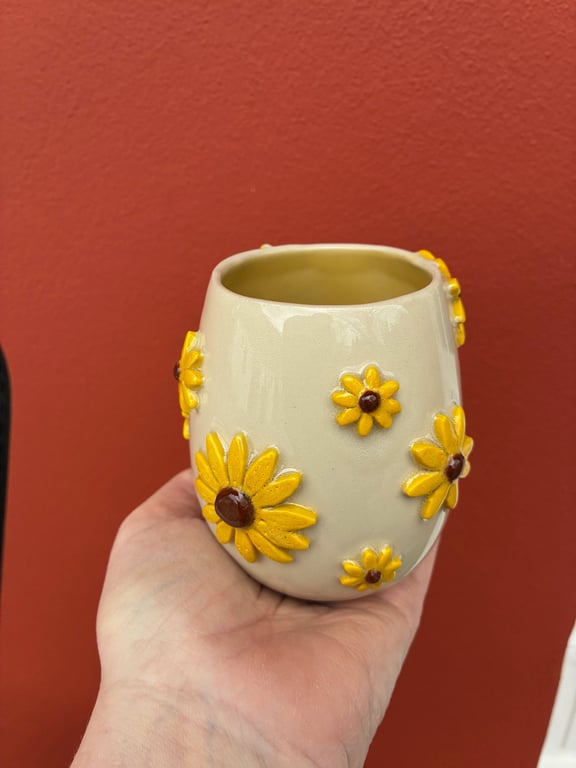 Hånd, der holder en beige keramikkop med gule solsikkemotiver mod en rød væg.