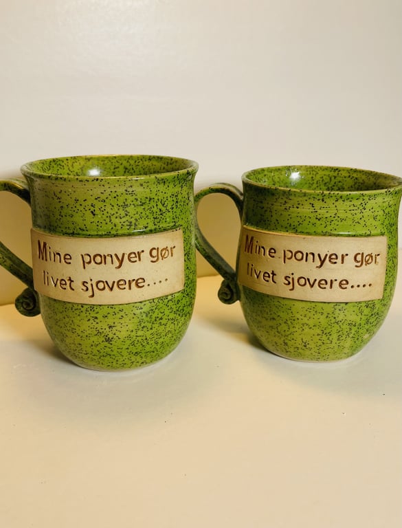 To grønne krus med sort prikmønster og hank, hvorpå teksten "Mine ponyer gør livet sjovere...." er skrevet på en beige rektangulær plade.