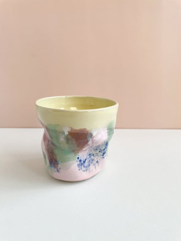 En lille, håndlavet keramik kop med farvede glasurer i pastelfarver som gul, grøn, pink, blå og brun på en hvid bordplade med en beige væg i baggrunden.