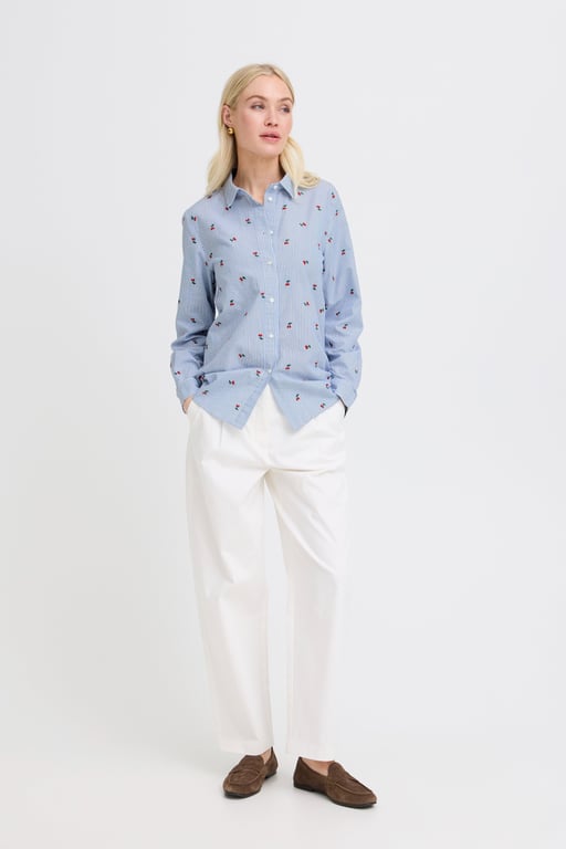 Kvinde iført lyseblå, langærmet skjorte med små mønstre og hvide bukser, står med hænderne i lommerne og ser til siden.