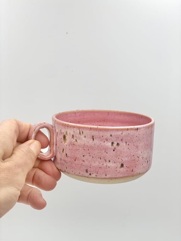 En hånd holder en lyserød keramisk kop med små mørke pletter mod en lys, neutral baggrund.