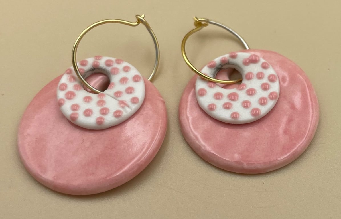 Øreringe af keramik med store, runde, lyserøde plader og mindre hvide cirkler med lyserøde prikker, hængende på gyldne øreringe.
