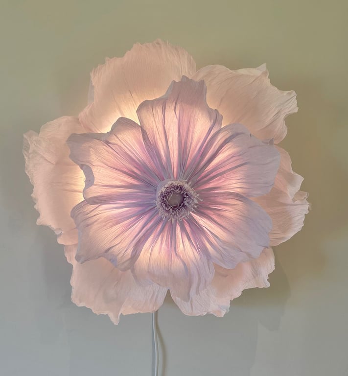 Blomstformet lampe med bløde, lyserøde kronblade, der lyser op mod en neutral væg.