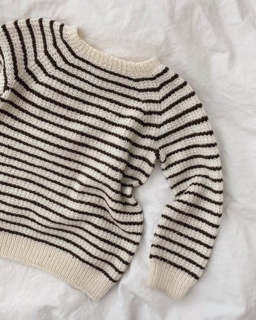 En strikket sweater med cremefarvet bund og sorte horisontale striber, lagt fladt på en hvid overflade. (1)