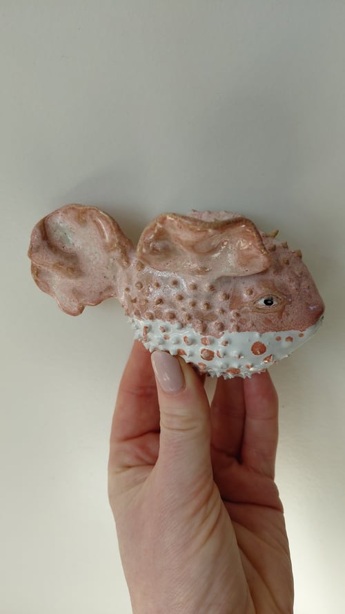 Hånd, der holder en lille, brunlig keramikfigur formet som en pigfisk med hvid og orange prikket mave. (1)