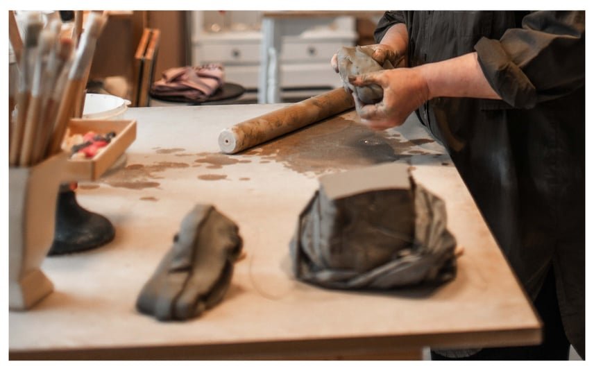 En person arbejder med ler ved et bord, hvor lerklumper og en kagerulle ligger.