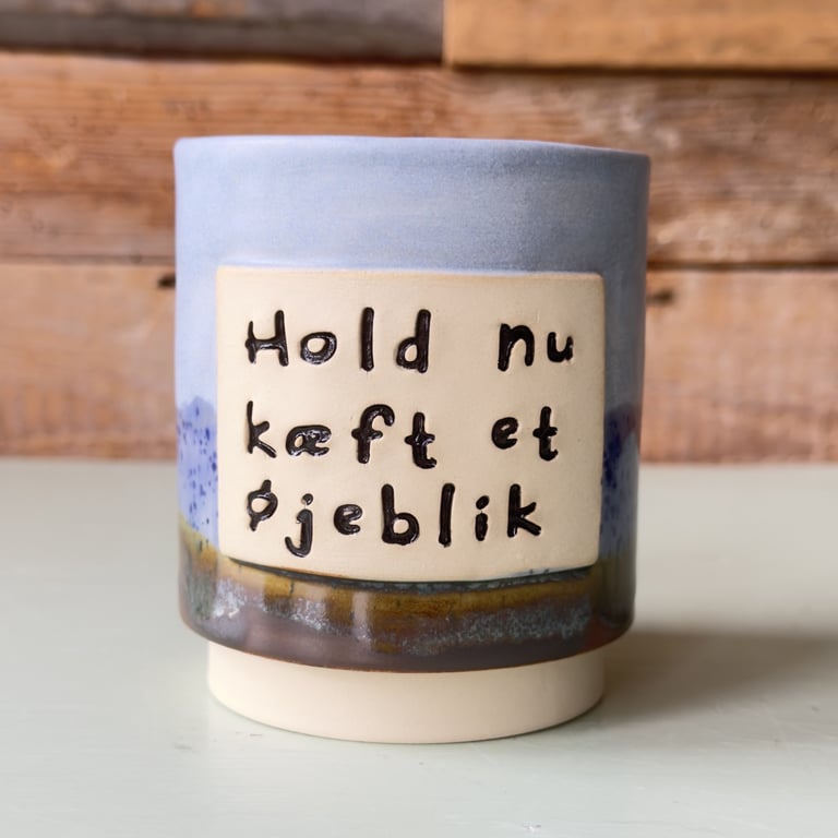Keramikkop med blå, grønne og brune nuancer med teksten "Hold nu kæft et øjeblik" skrevet på dansk.