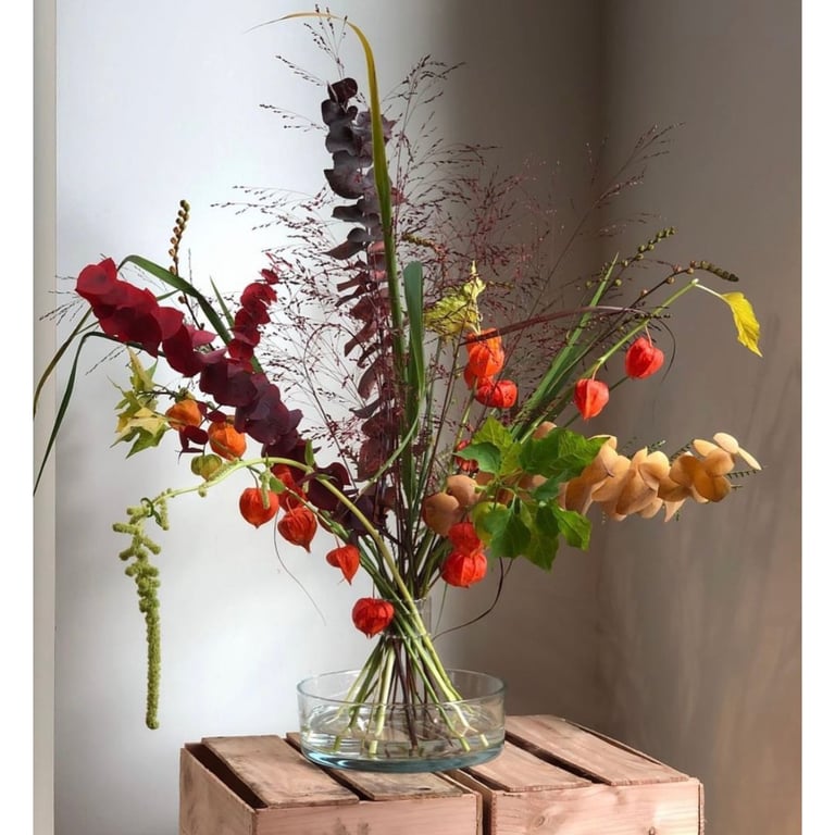 Bukket med tørrede og friske efterårsblomster i glasvase på træbord, bl.a. med orangerøde lygteblomster, brune og grønne blade samt høje græsstrå.