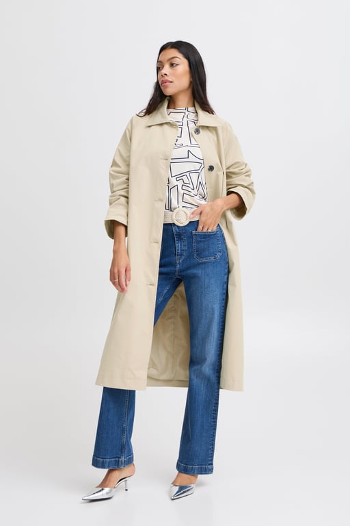 Kvinde iført beige trenchcoat, mønstret top, blå jeans og sølvfarvede hæle, stående mod hvid baggrund.