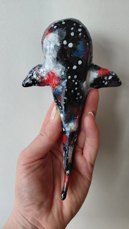 Hånd, der holder en lille, håndlavet figur formet som en hval med sort, rød, blå og hvid farvet mønster.