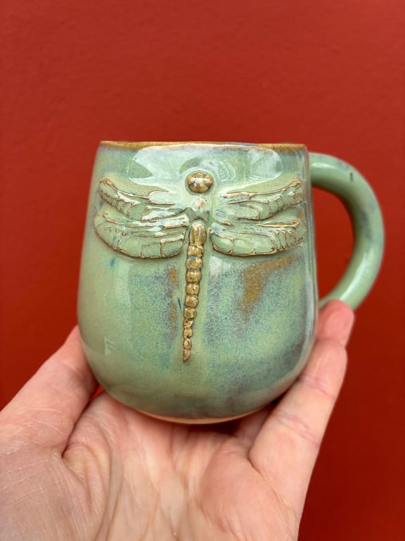 Hånd, der holder en grøn keramikkop med et relief af en guldfarvet guldsmed foran en rød væg.