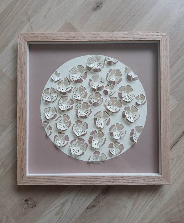 Billedkunst bestående af udskårne papirblomster arrangeret i en rund form med lysebrun baggrund og lyst træramme.