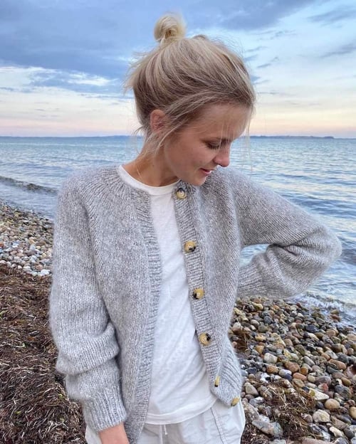 Kvinde i grå strikket cardigan og hvid t-shirt står på en stenstrand med havet i baggrunden ved solnedgang. (1)