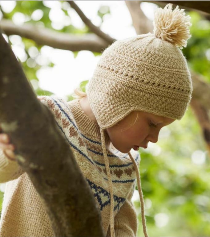 Barn med beige strikhue med dusk og mønstret sweater holder fast i et træ i en udendørs grøn baggrund.