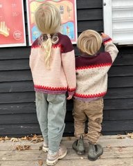 To børn iført strikkede sweater står med ryggen til foran en sort trævæg med plakater. (3)