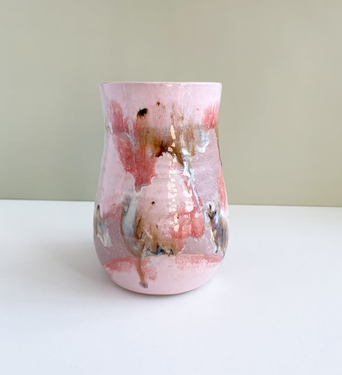 Lyserød keramikvase med abstrakte brune, hvide og sorte malingspletter stående på en hvid overflade foran en beige væg.