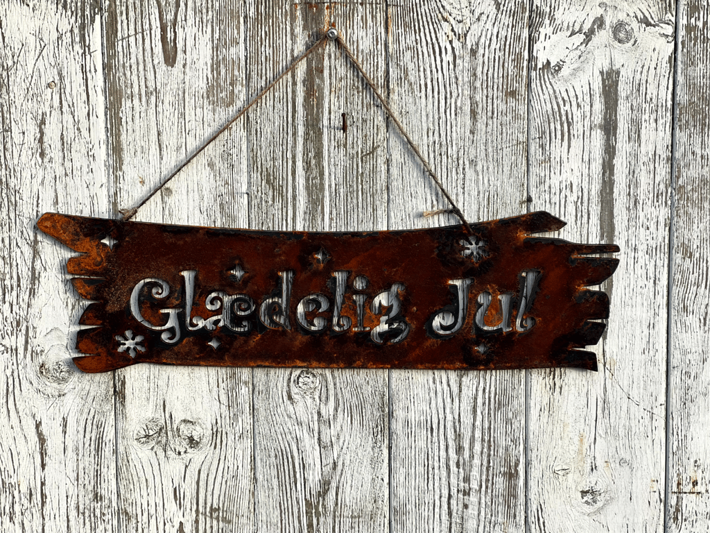 Rustent metalophæng med teksten "Glædelig Jul" hængende på en hvidmalet trævæg.