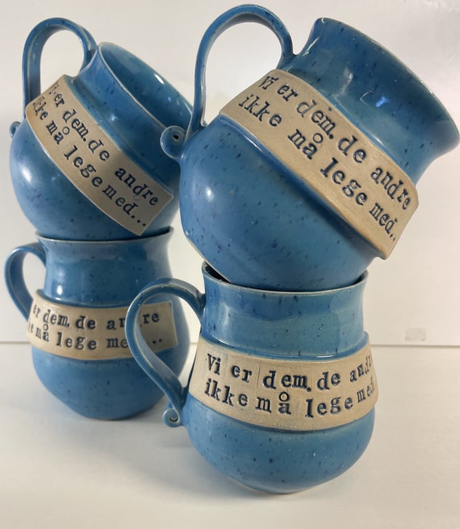 Billede af fire blå keramikkrus med hank, stakket i to bunker, med teksten "Vi er dem, de andre ikke må lege med" på en beige stribe rundt om hvert krus.