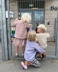 Tre små børn med lyst hår, hvoraf to står og kigger gennem et metalgitter, mens det tredje barn knæler og holder om det ene barns ben. (2)