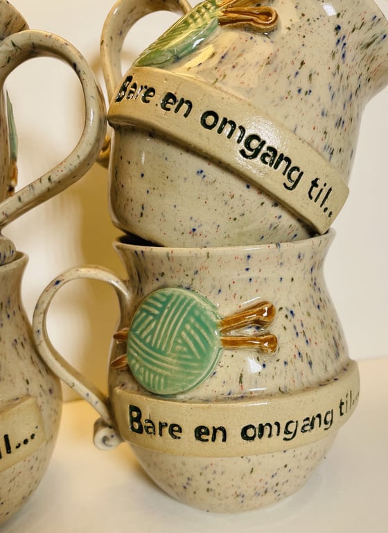 Stentøjskrus med grøn kugle af garn og pinde som pynt, samt teksten "Bare en omgang til..." skrevet i sort.